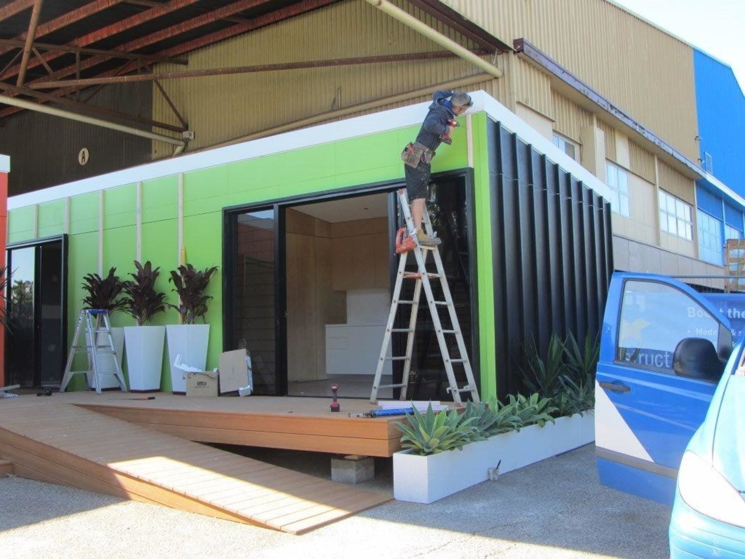 The same module close to complete — lime green painted panels with a white reveal above, the black battened end wall at close centres, aluminium sliding doors onto a composite timber deck with a ramp, planters of cordylines and agaves along the front, and a carpenter on a ladder fixing the final trim at the corner