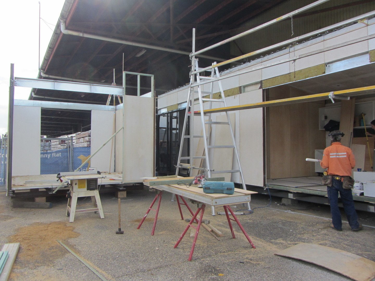 Two modules under construction in the fabrication yard — steel chassis on blocks, wall panels standing with insulation still exposed at the head, a saw bench and trestles set up on the gravel, and a carpenter working at the open end of the far module