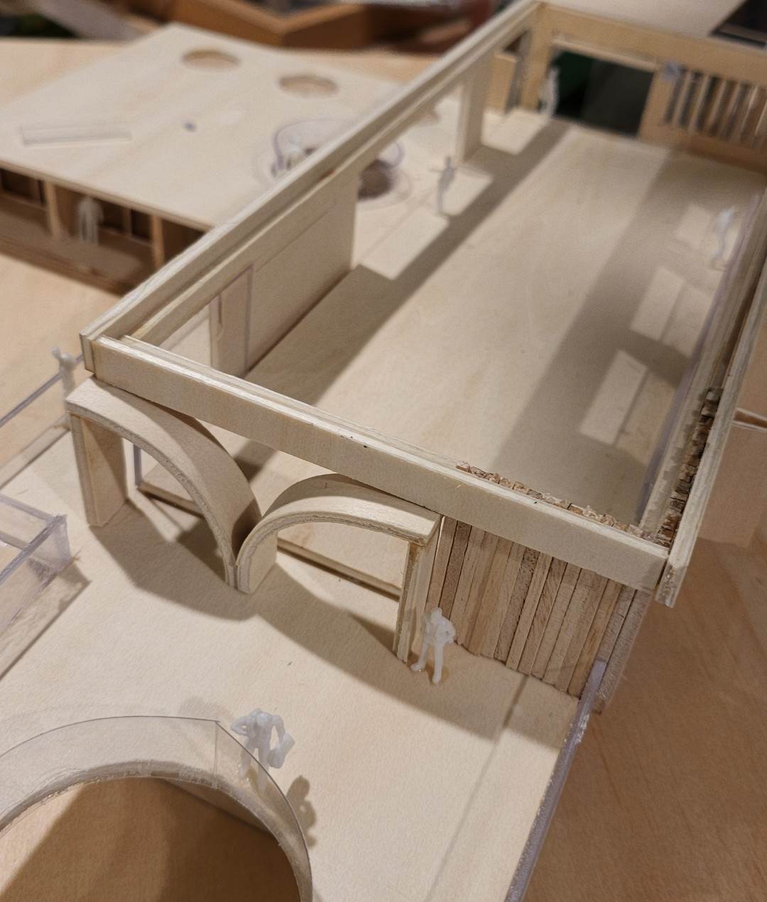 Close view of the physical model showing the two arched openings beneath the long upper beam, beside a wall of fine timber slats, with small white figures standing at the entry
