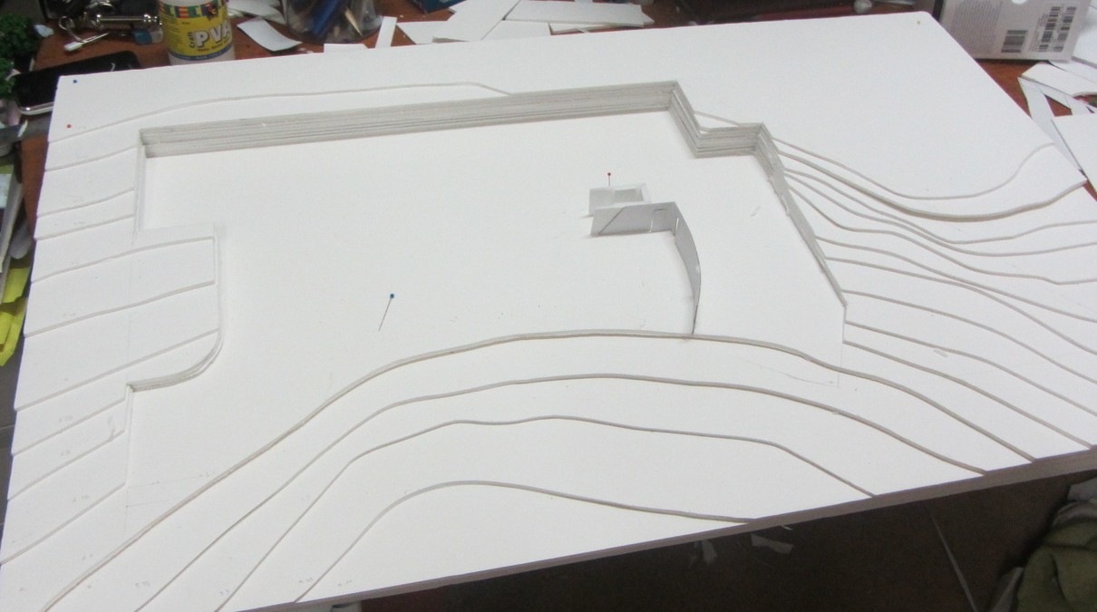 The contour model on the workbench &mdash; stacked foamboard layers stepping across the site to show the existing fall, with the building footprint cut into the terraced ground