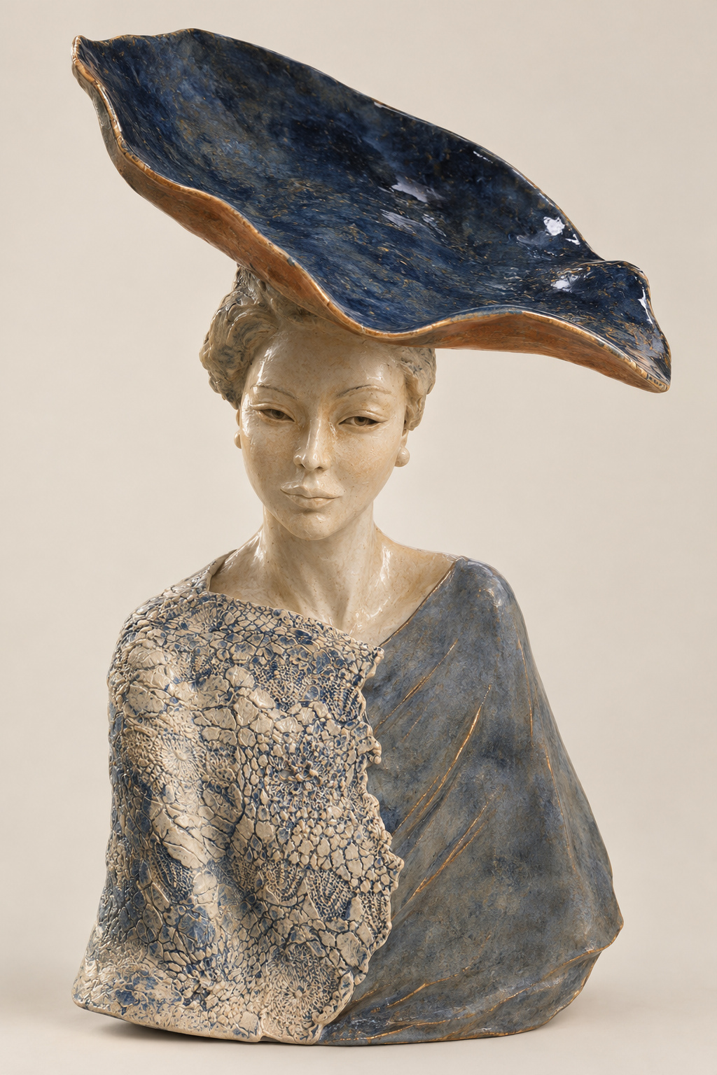 The same bust after glazing and firing — the wide brim now a deep, glossy blue flecked with copper, the face in cream glaze, and the lace-textured shoulder picked out in blue against a soft blue-grey drape