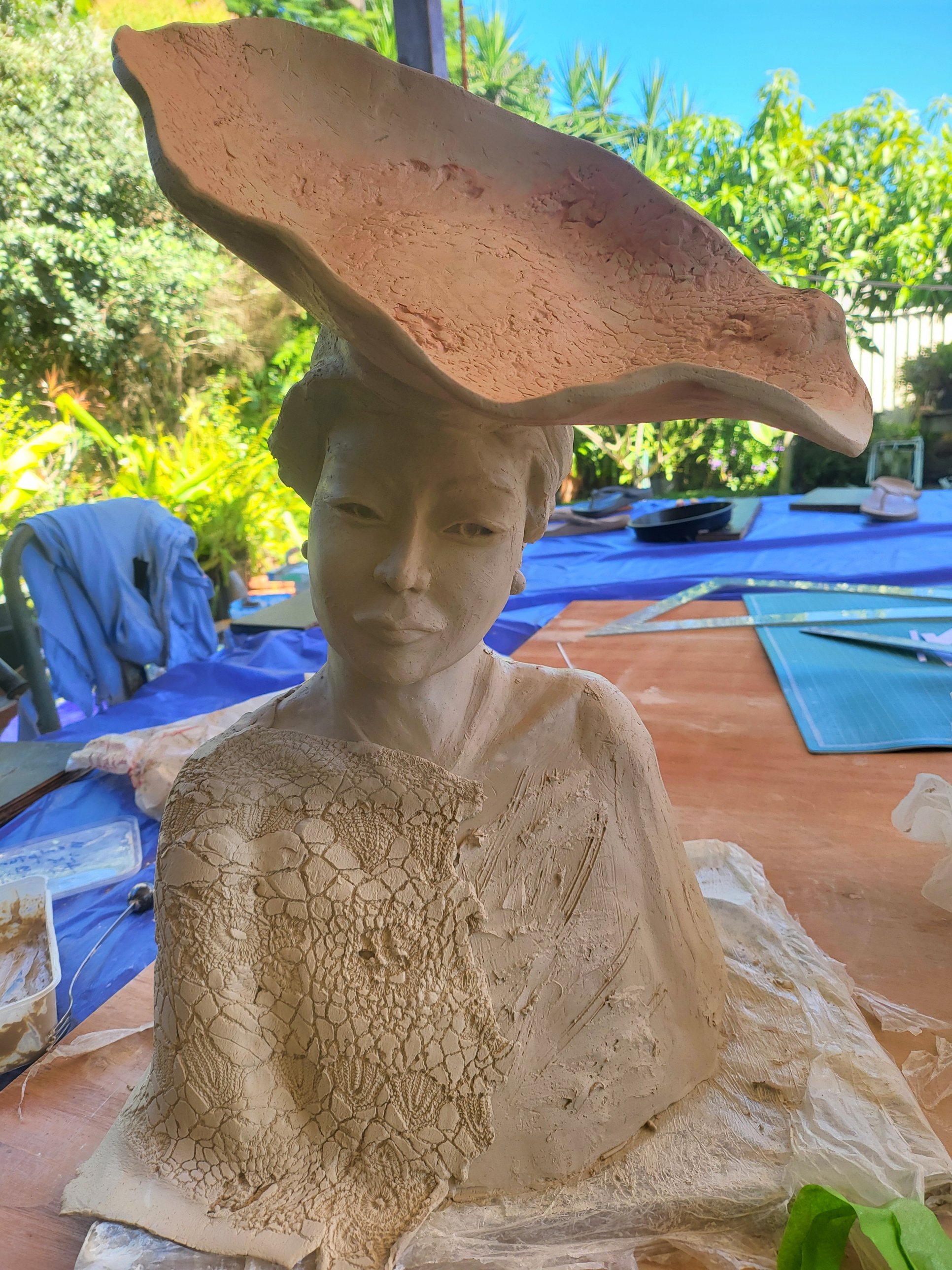 A portrait bust in greenware outdoors — a wide undulating clay brim tilted above the head, a calm modelled face, and one shoulder draped in a cloth pressed all over with lace texture