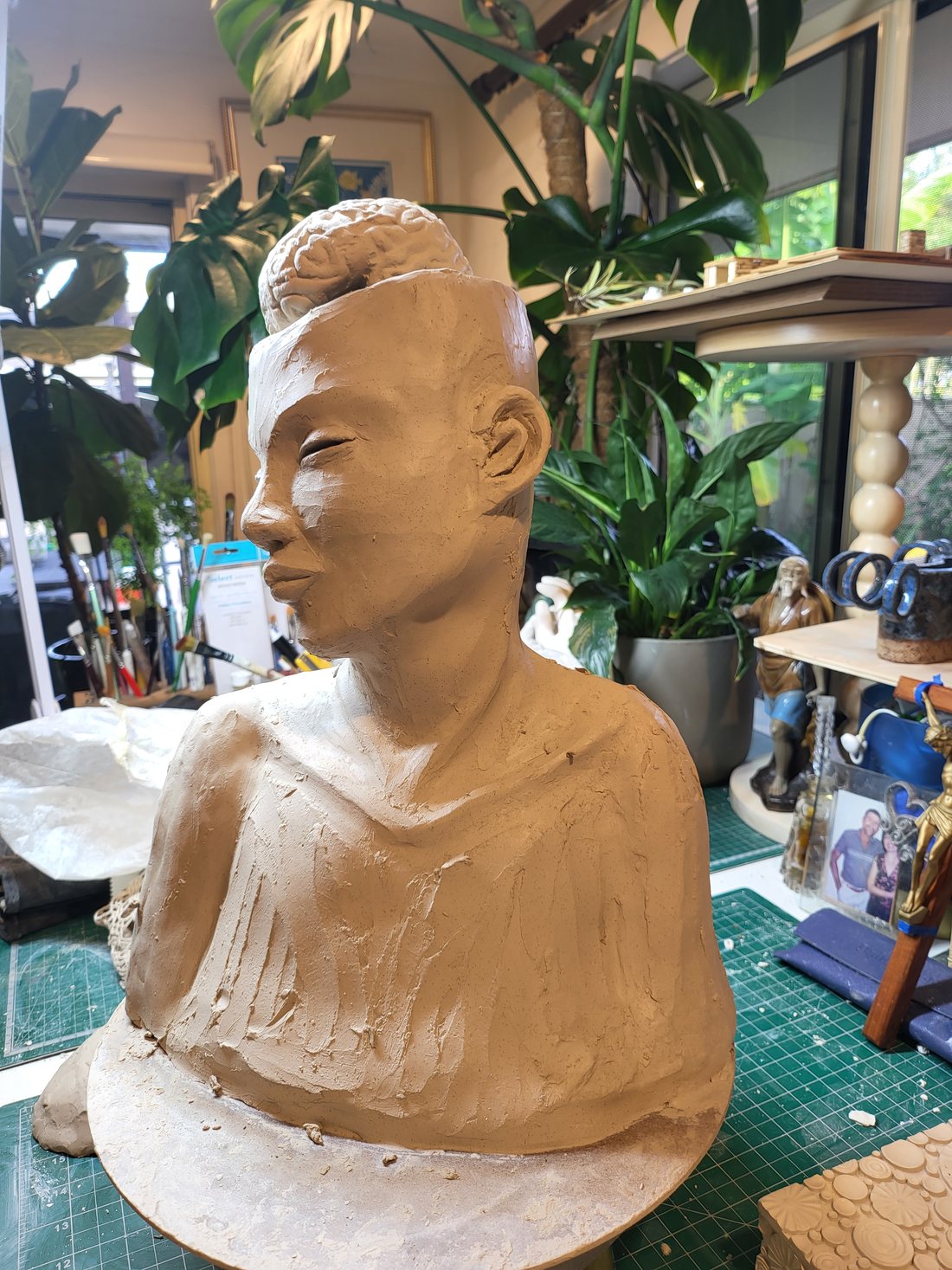 A portrait bust in greenware on the studio turntable, seen from the side — a sculpted brain rising from the open top of the head, eyes softly closed, the shoulders and chest roughly worked with the marks of the modelling tools still visible