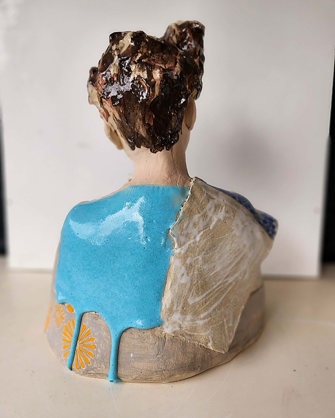 The same glazed portrait bust, back view — a glossy turquoise blue glaze pooling and dripping down the back over pale stoneware, with gold fan motifs at the base