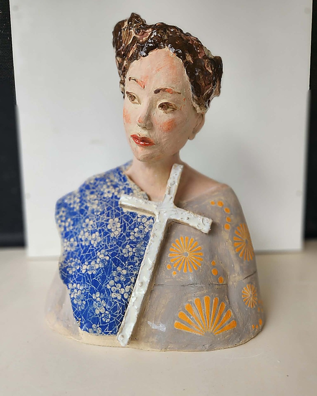 A glazed portrait bust, front view — dark modelled hair, softly coloured face, the shoulders draped in a cape of deep blue blossom pattern with a raised white cross and gold fan motifs