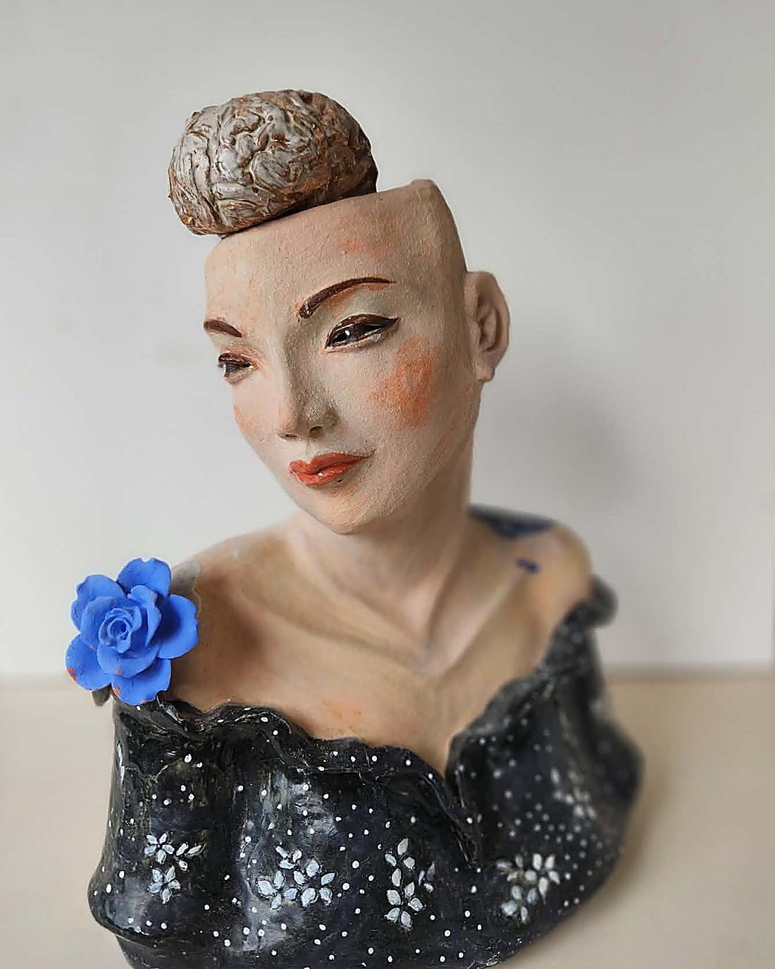 A glazed portrait bust, front view — a sculpted brain sitting where the hair would be, a downcast painted face with blushed cheeks and red-orange lips, a blue ceramic rose on the shoulder, and a black dress with white dotted floral glaze detail