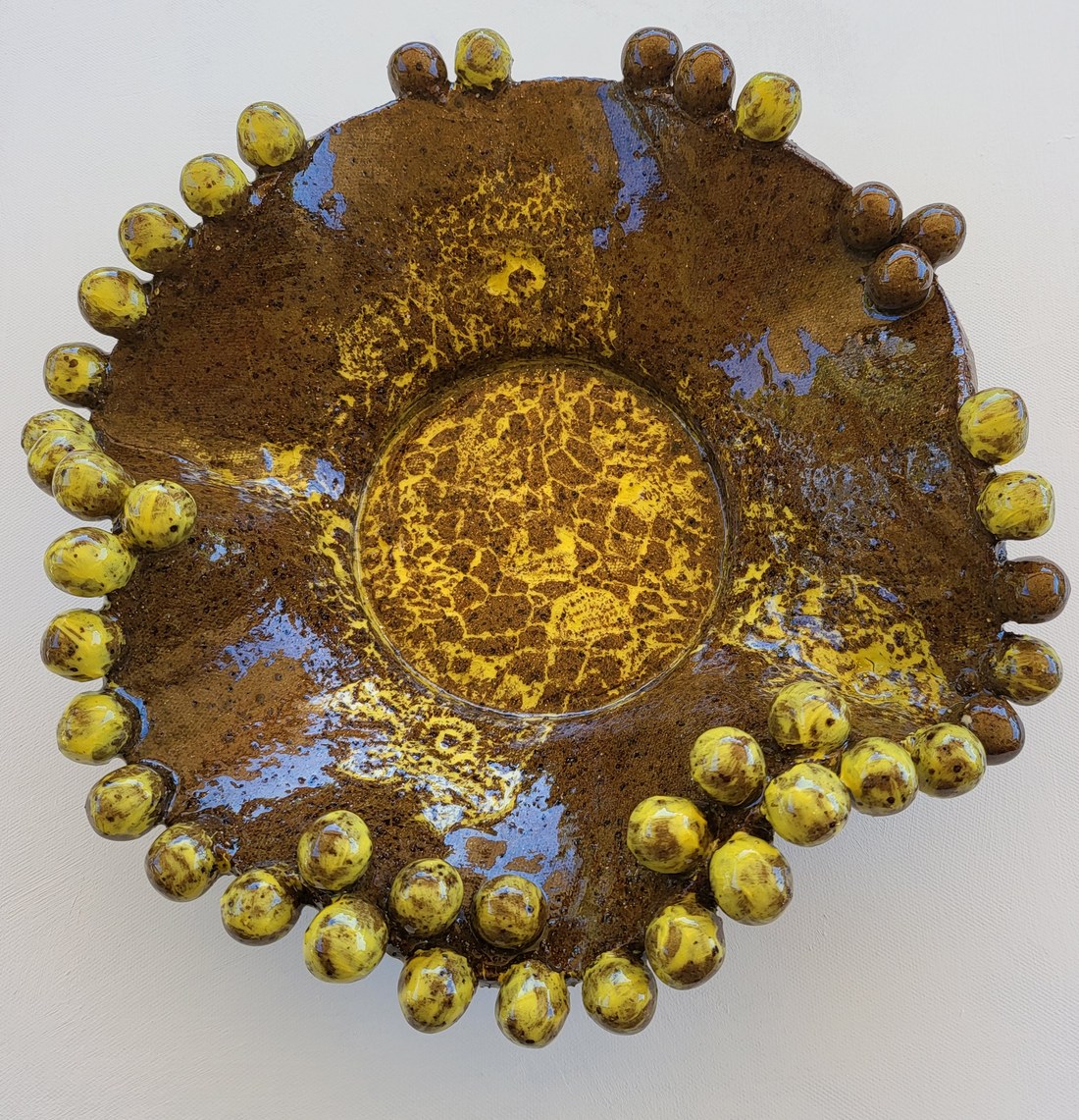A shallow round plate in a glossy treacle-brown glaze with lilac pooling, a lace-textured centre showing through in bright yellow, and a rim built from a close ring of clay balls glazed yellow and brown