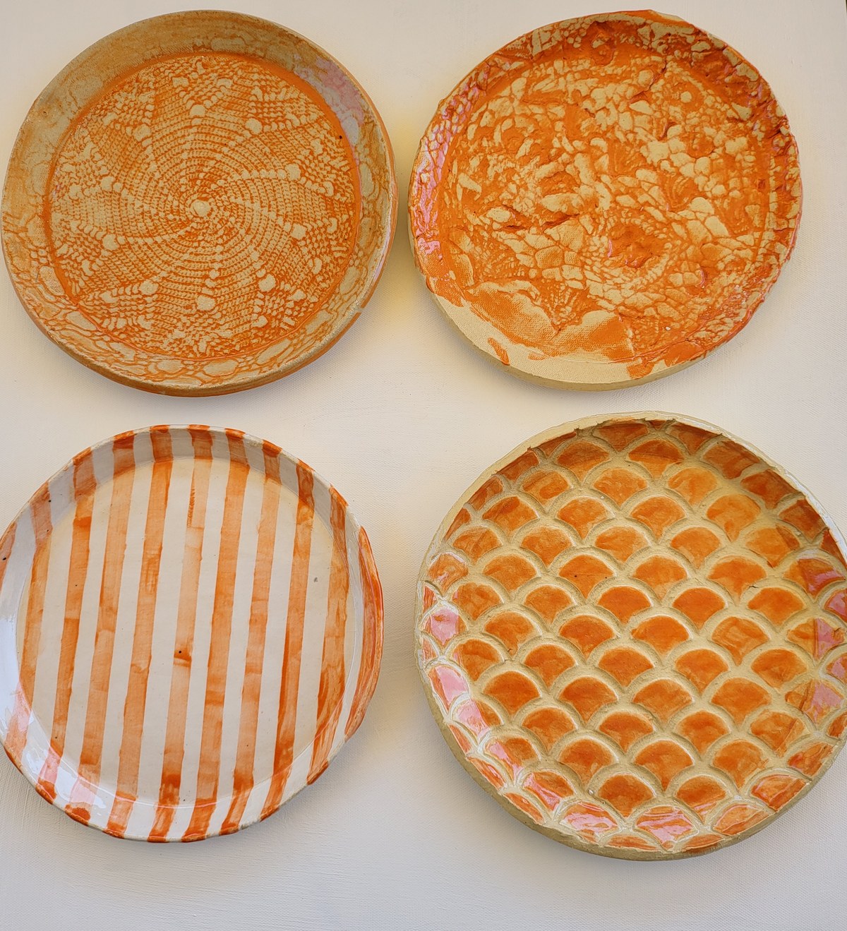 Four round plates laid out in a square — the top two in orange slip with lace and doily patterns carved back through to the pale clay, the lower left painted in orange and white stripes, and the lower right an embossed fish-scale relief with orange underglaze in the recesses