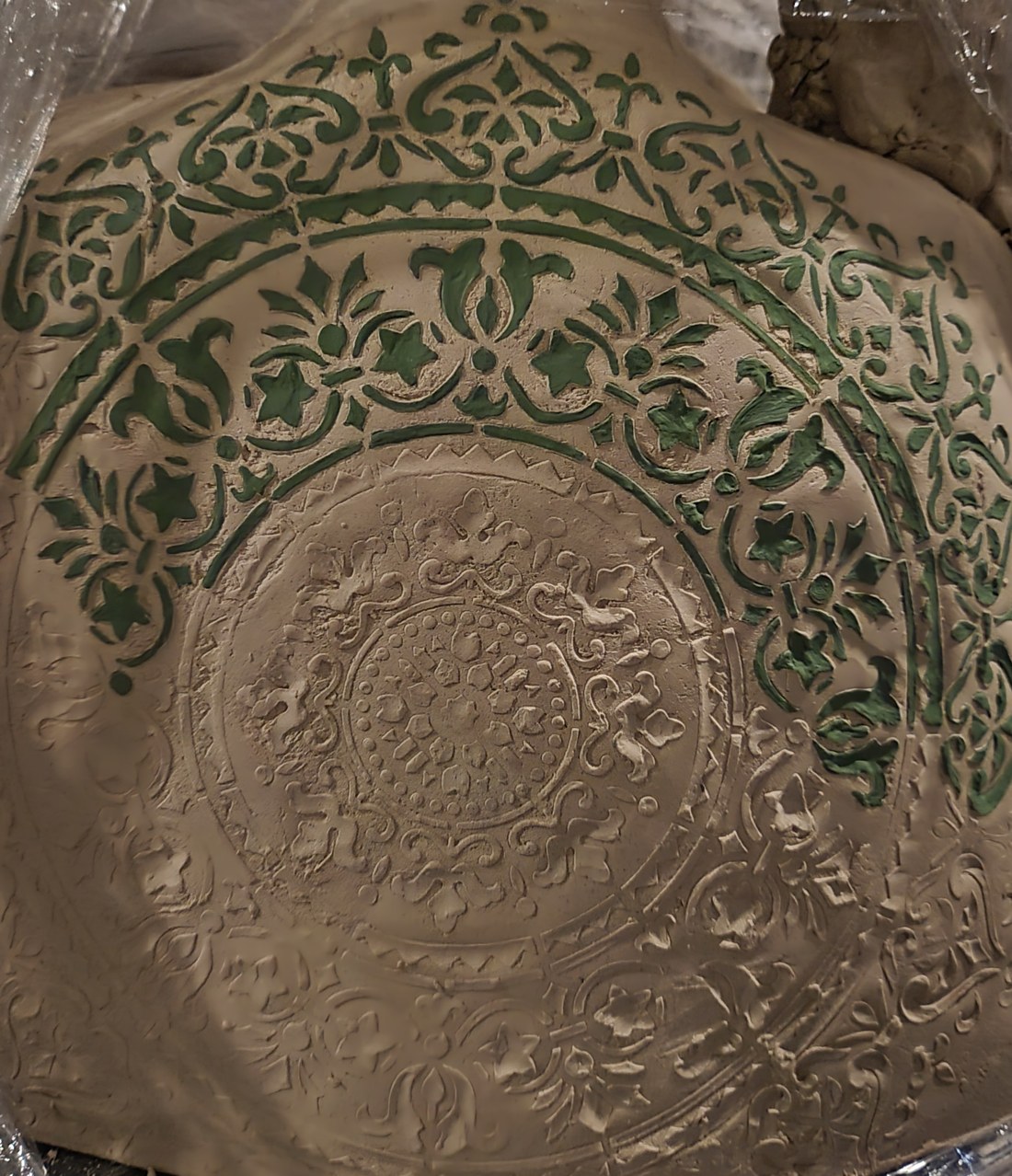 A square slab of pale unfired clay, a lace-like stencil pattern pressed directly into the damp surface — an embossed medallion at the centre and a border of scrolling foliage, with green underglaze brushed into the pattern across the upper half