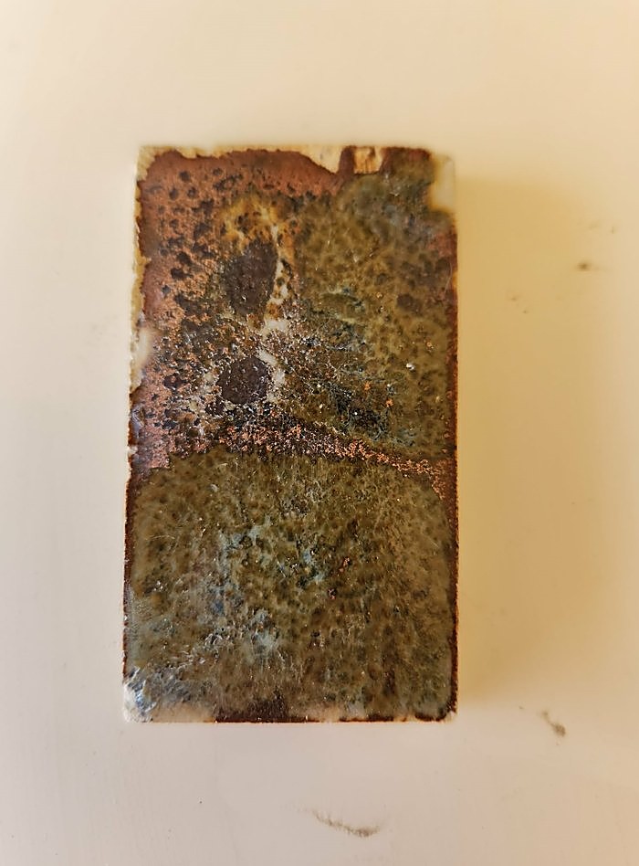 A test tile layered Bailey Red, then Selsor White, then clear glaze — fired to a dense mottled olive, copper and rust surface