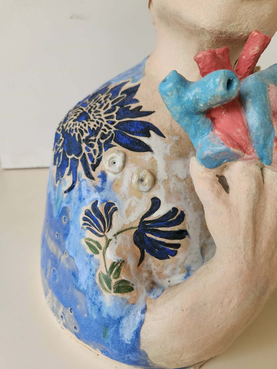 The same bust after glazing and firing — the cobalt blue flowers now deep and glossy, a mottled blue glaze across the shoulder and body, and the heart at the neck in bright blue and coral pink