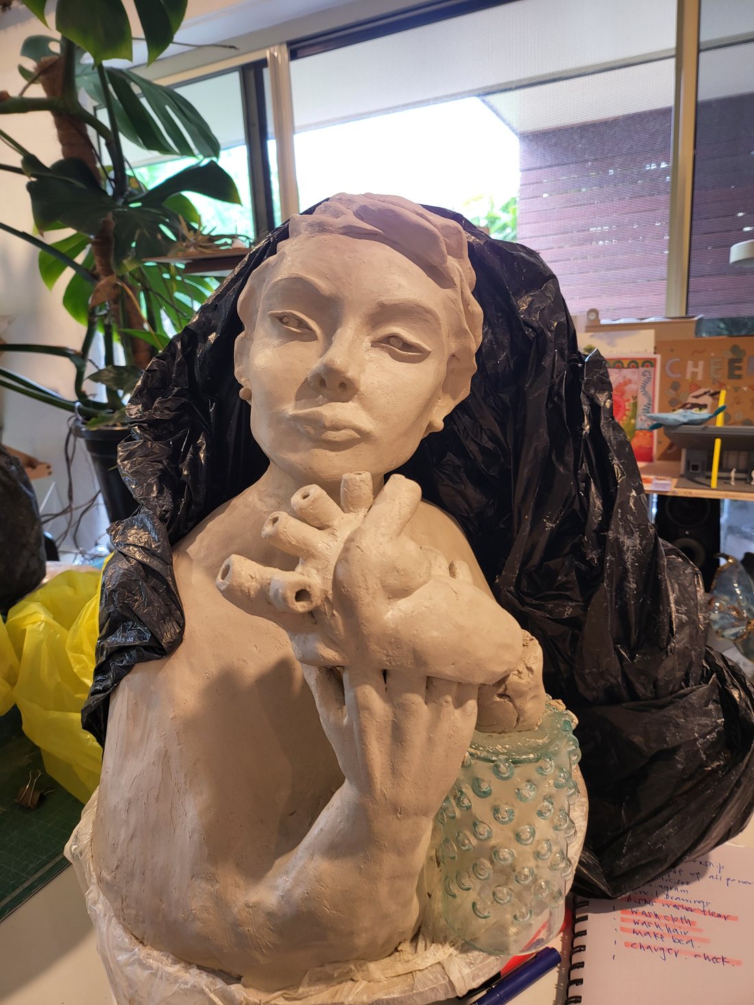 A female bust in pale greenware, front view — short sculpted hair, a calm direct gaze, and both hands raised to the chest holding an anatomical heart with its open vessels