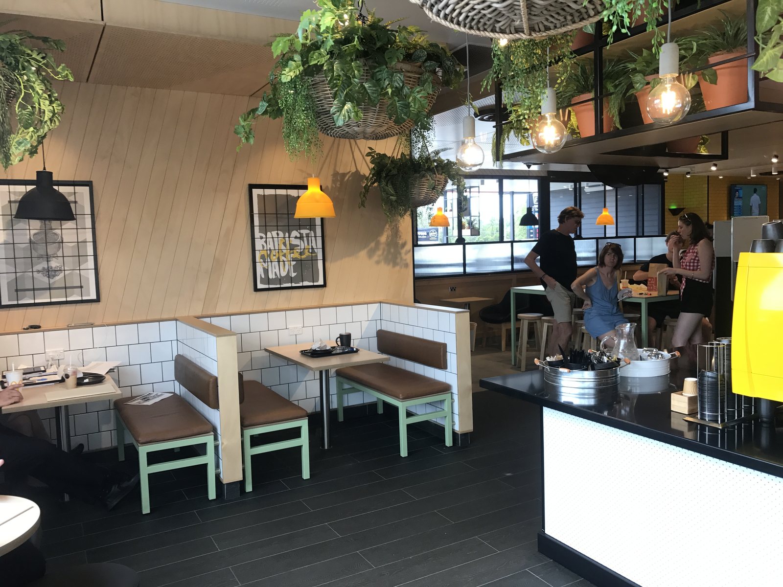 The completed banquette area &mdash; angled pale timber wall, black and yellow pendants, white tiled low walls with timber ends, brown upholstered benches on pale green steel frames, and hanging planting overhead
