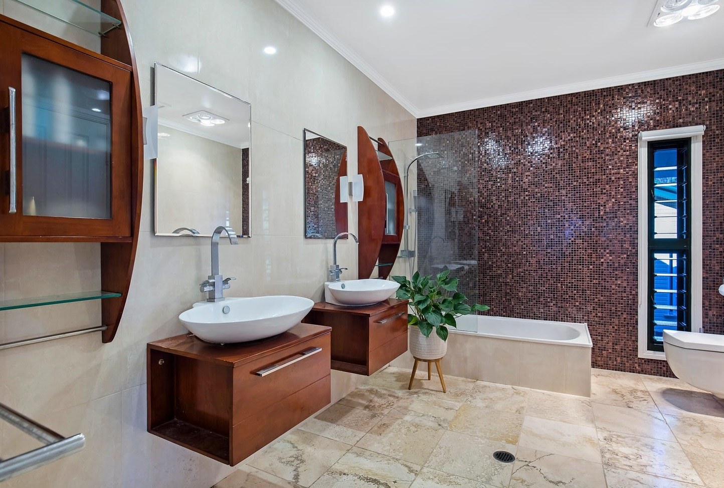 The completed ensuite — twin timber vanities with white vessel basins, a bath against a deep chocolate glass mosaic wall and travertine flooring