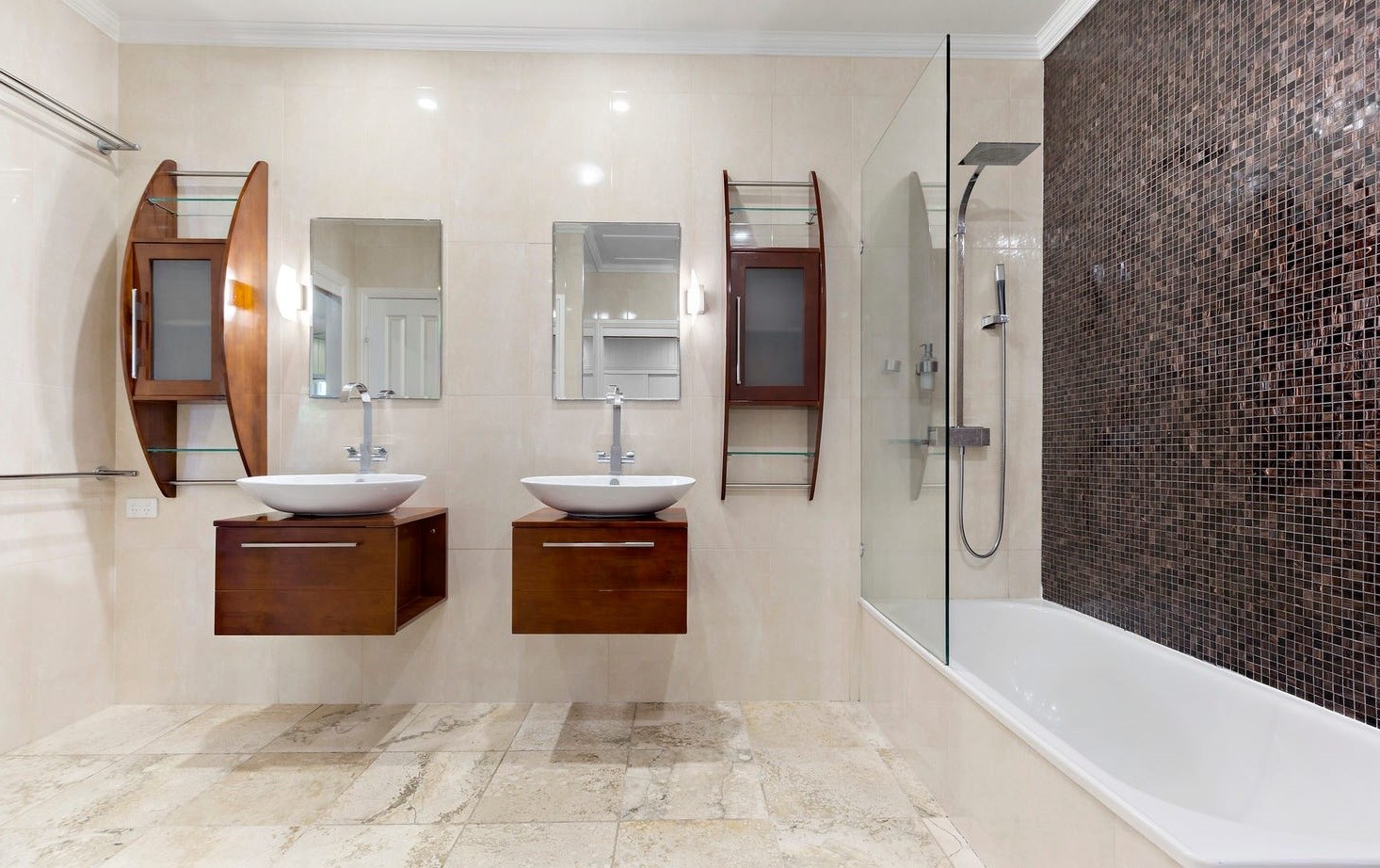 The twin vanities seen square on &mdash; two timber units with white vessel basins and wall-mounted mixers, a mirror above each, curved timber shelving units either side, and the glazed shower over the bath against the mosaic wall