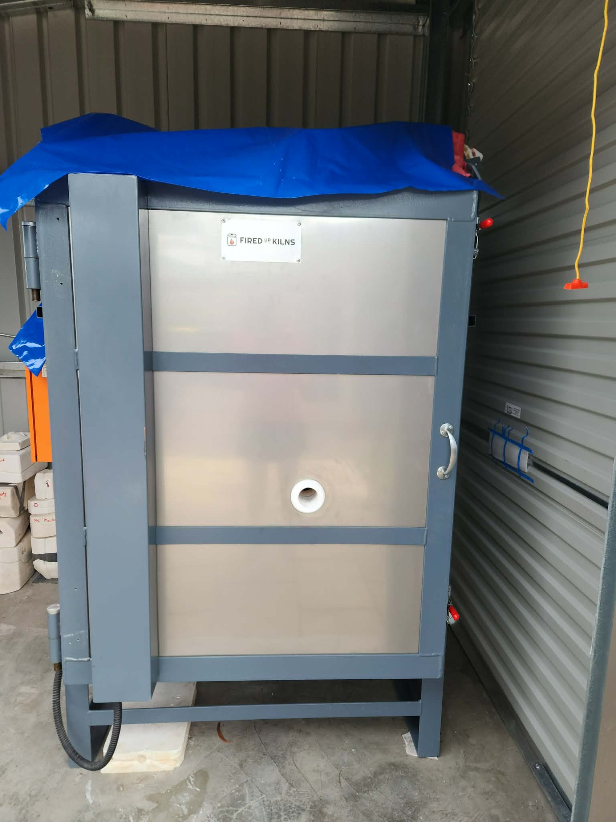 A new electric kiln in its shed, still under a blue tarp, an orange control cabinet standing beside it