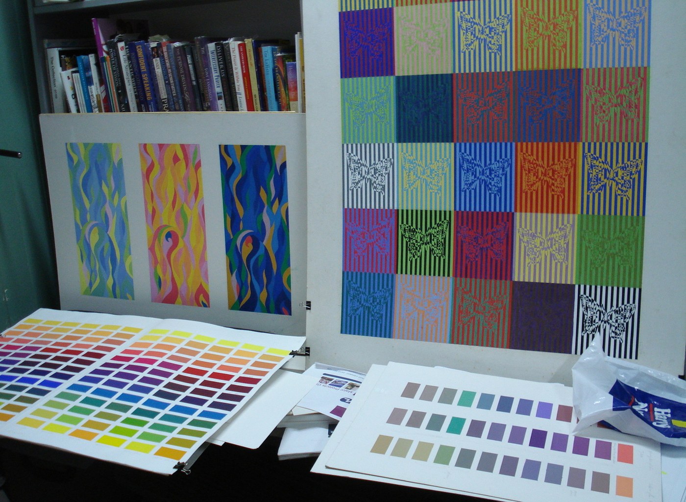 The Butterflies grid pinned up on a studio display board beside a triptych of flowing colour studies, with large printed colour charts and painted swatch sheets laid out across the bench below and a shelf of design books above