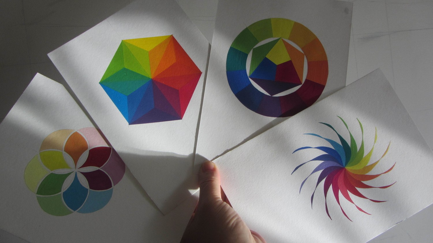 Four colour wheel studies fanned out and held in one hand in raking daylight &mdash; a six-sided wheel divided into triangular facets, a twelve-part ring around an inner triad, a rosette of overlapping translucent circles, and a pinwheel of tapering petals running through the full spectrum