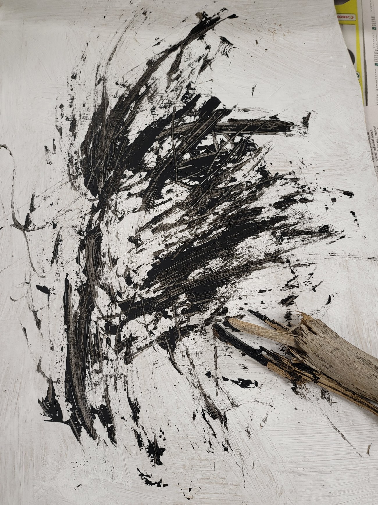 A gestural black work on a white ground &mdash; strokes dragged and struck with a tree branch, fanning out from a dense knot at the centre into flicked marks and spatter, the weathered branch still resting on the sheet