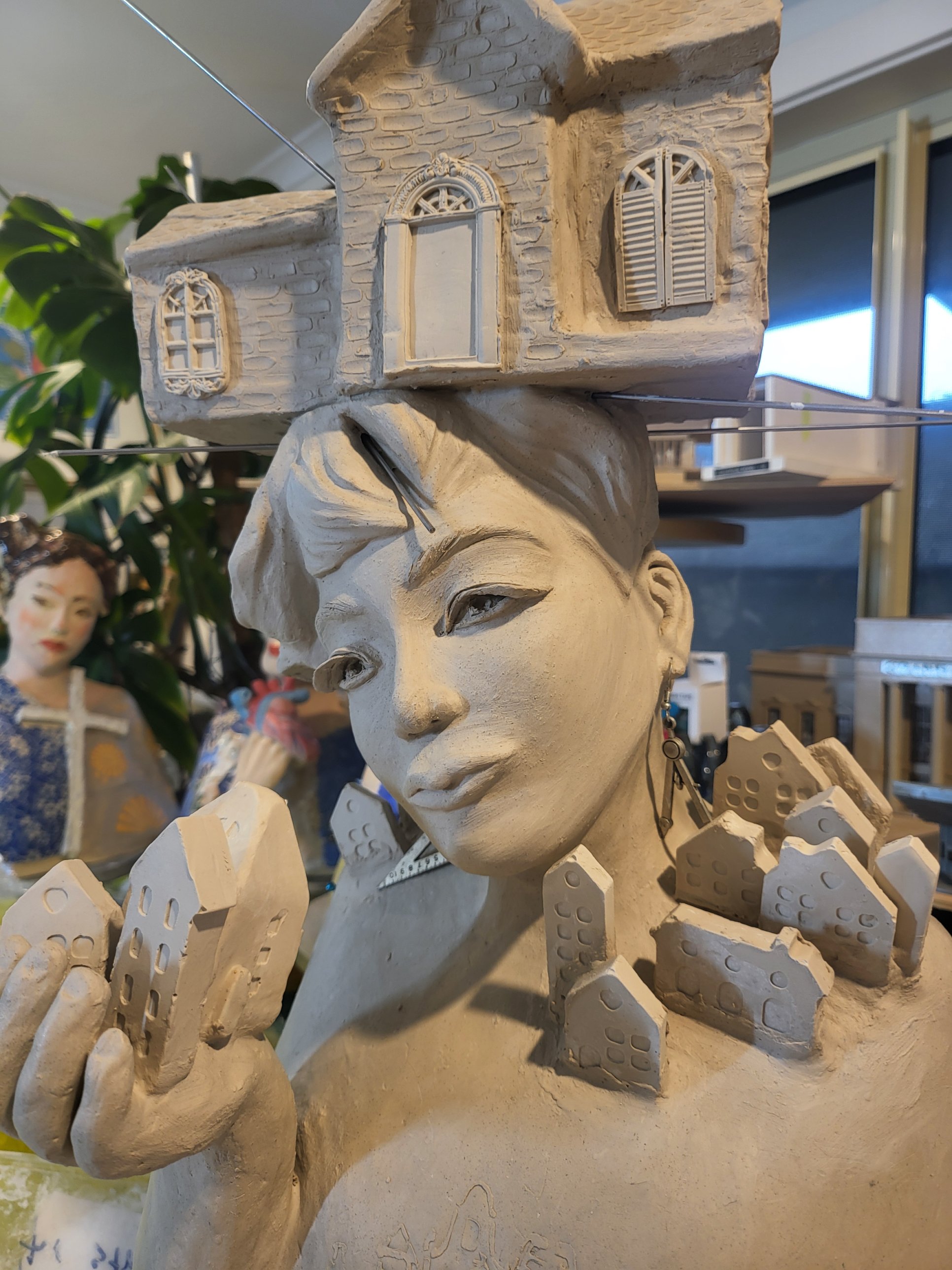The Architect — a house resting above her head, small houses held in her hand and rising from her shoulder
