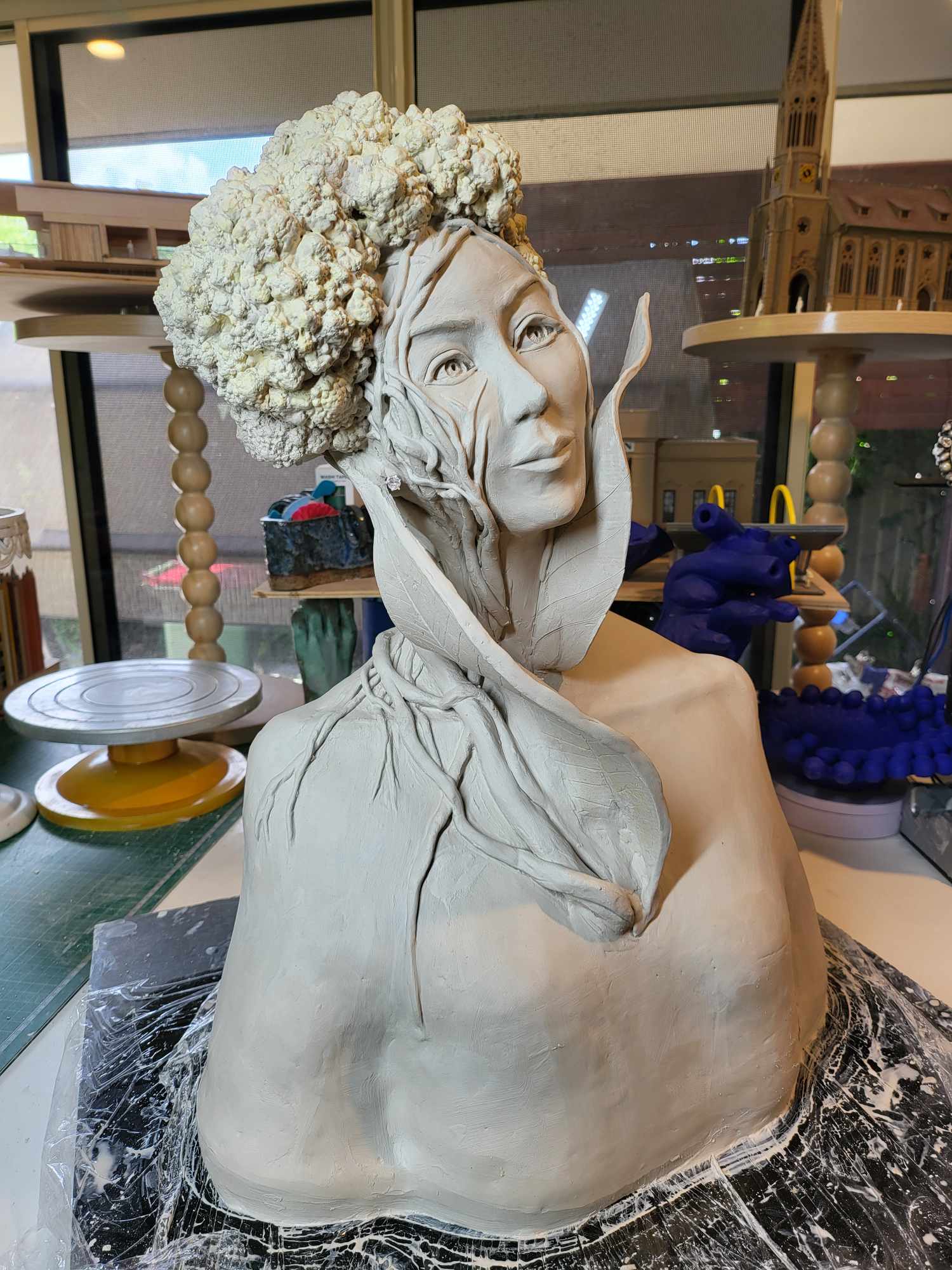 The Horticulturist in greenware beside a real cauliflower on a stand &mdash; the sculpted hair built as dense cauliflower florets, leaf forms rising at the shoulders and collar, the body still wrapped to keep the clay damp