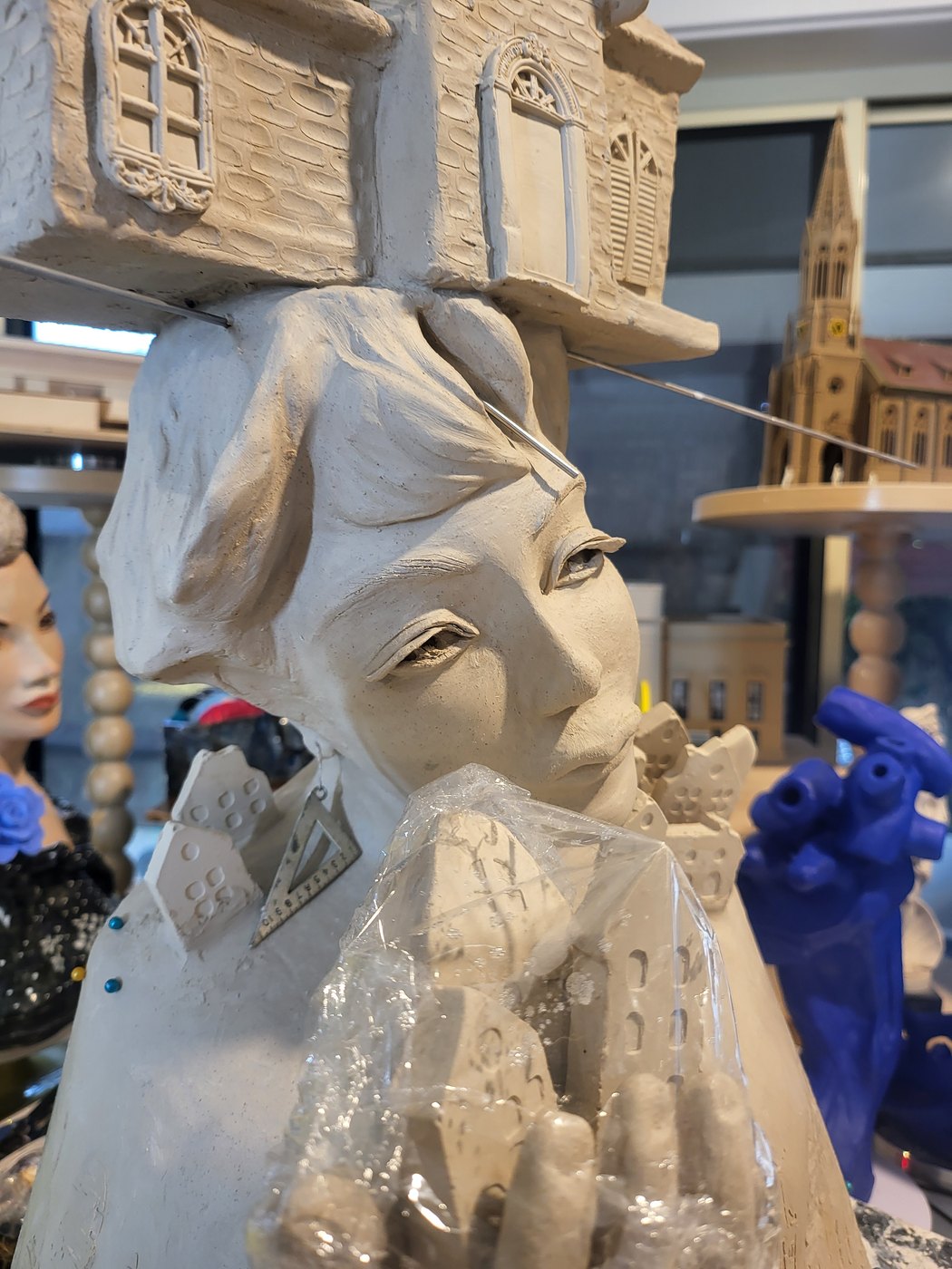 Close view of the Architect in greenware — her face turned slightly downward beneath a detailed house with arched windows and shutters, a metal set-square hanging from her ear, and small houses rising from her shoulder and held in her hand under protective plastic