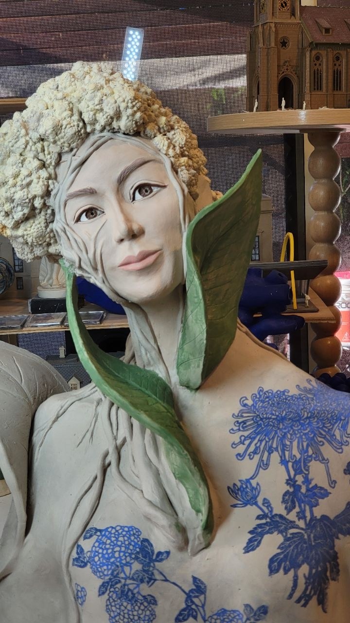 The Horticulturist close to the face &mdash; cauliflower hair above, fine roots running from the cheek down the neck, two green leaves rising on either side of the chin, brown painted eyes and soft pink lips, and blue botanical transfers across the chest