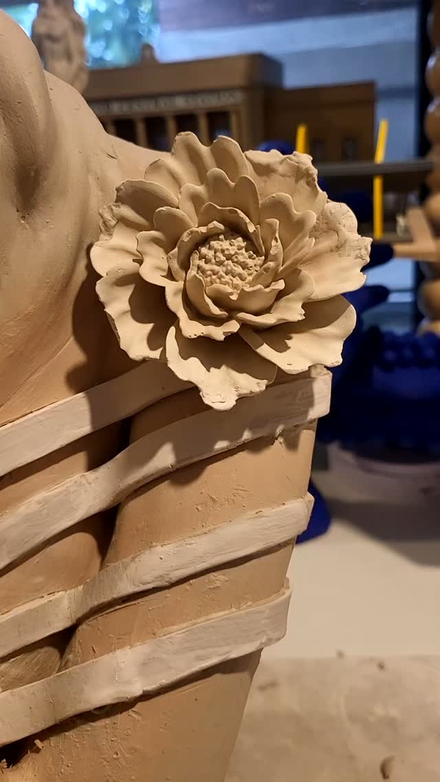 Close view of the shoulder &mdash; a sculpted peony with layered, ruffled petals and a textured centre, sitting above the flowing strips of clay that wrap the bodice