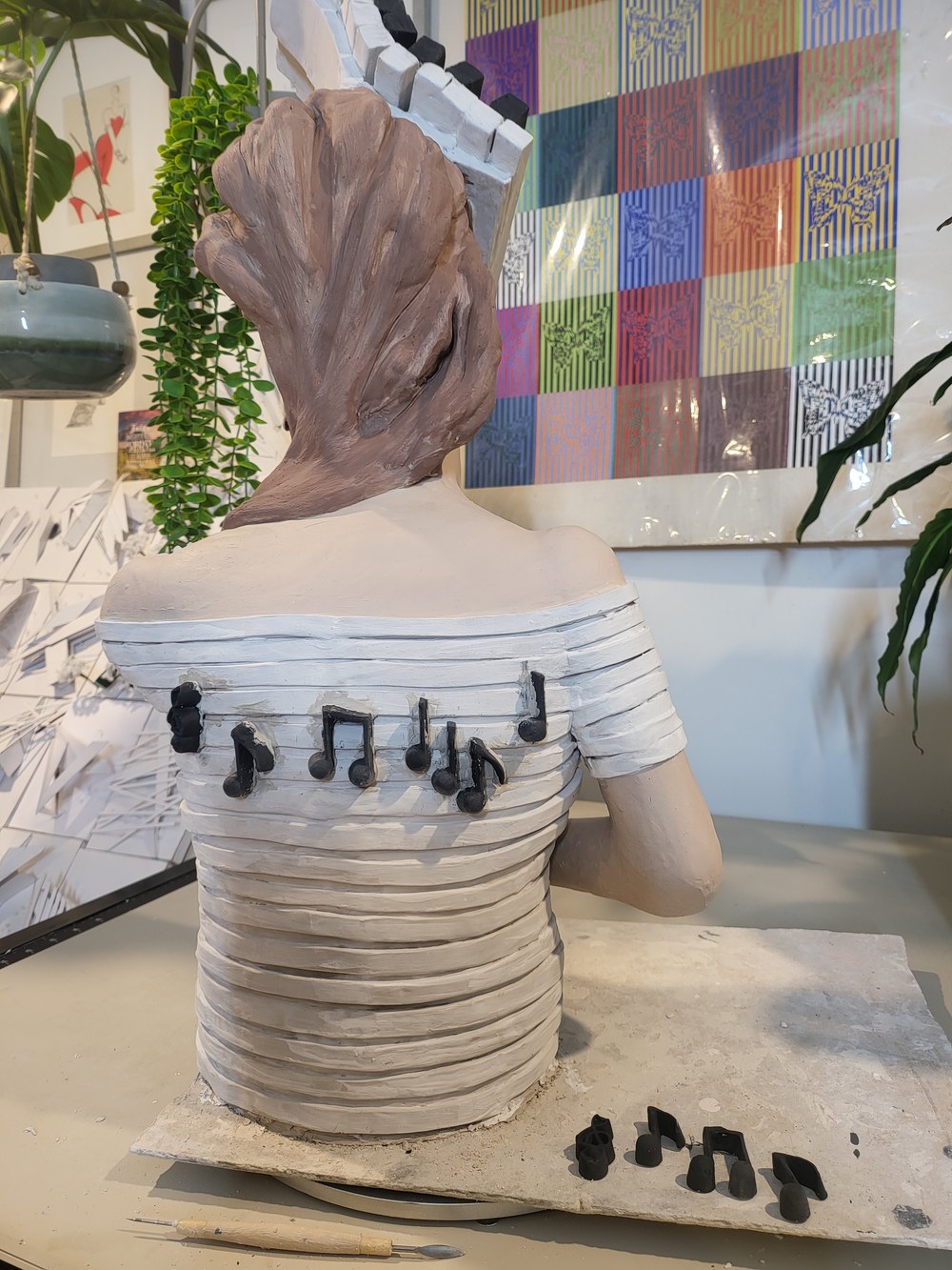 The back of the finished figure &mdash; hair coiled up off the neck, the bodice built from horizontal strips of clay, and a run of black musical notes laid across the upper back, with more notes waiting on the board beside her