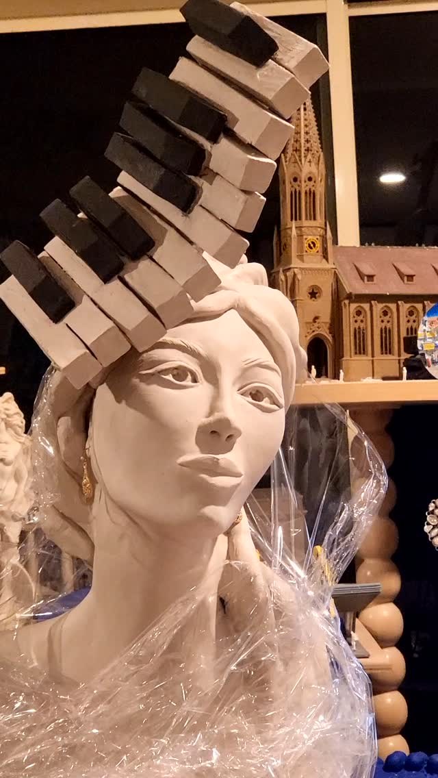 The face in greenware, lifted and calm, with a gold drop earring, the black and white piano keys rising in a stepped arc from one side of the head