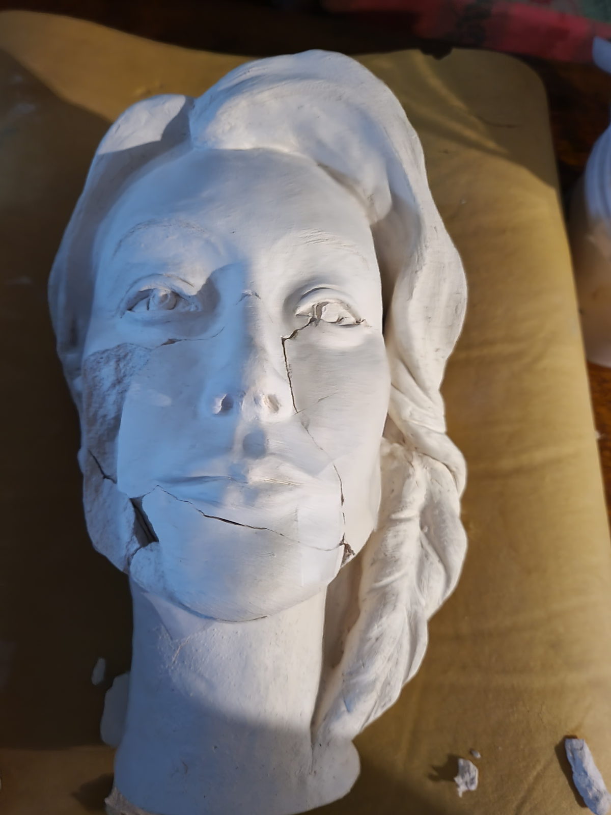 A bisque-fired portrait head in pale clay, lying on a cloth, with clear cracks running down one cheek and across the chin and jaw