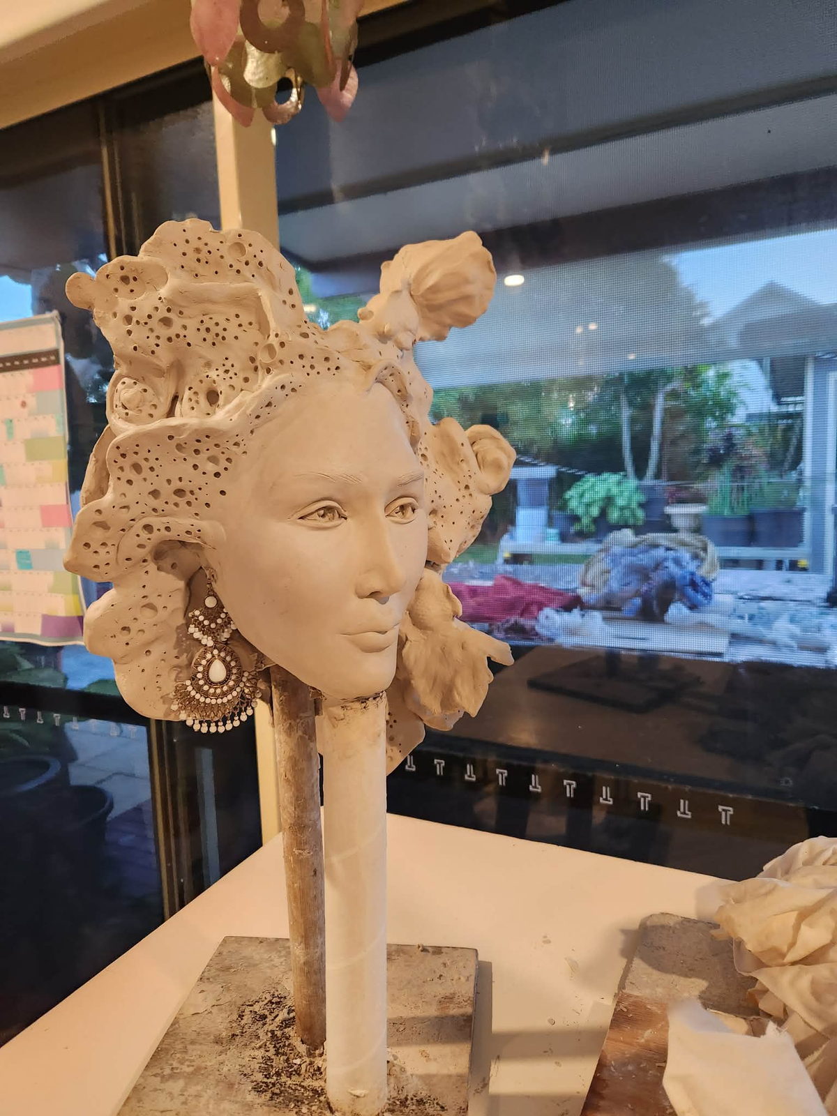 A finished portrait sculpture of a woman in unfired clay, framed by an elaborate perforated headdress, wearing a beaded chandelier earring
