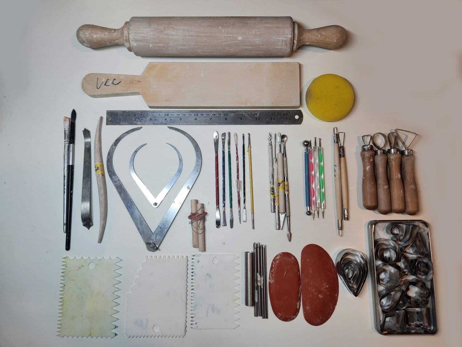 A fuller set of ceramic tools laid out &mdash; a rolling pin and wooden paddle, brushes, a metal rib, calipers, a steel ruler, fine modelling and detailing tools, ball and dotting tools, wire loop tools with wooden handles, serrated plastic scrapers, red kidney ribs, a sponge and a tin of small cutters