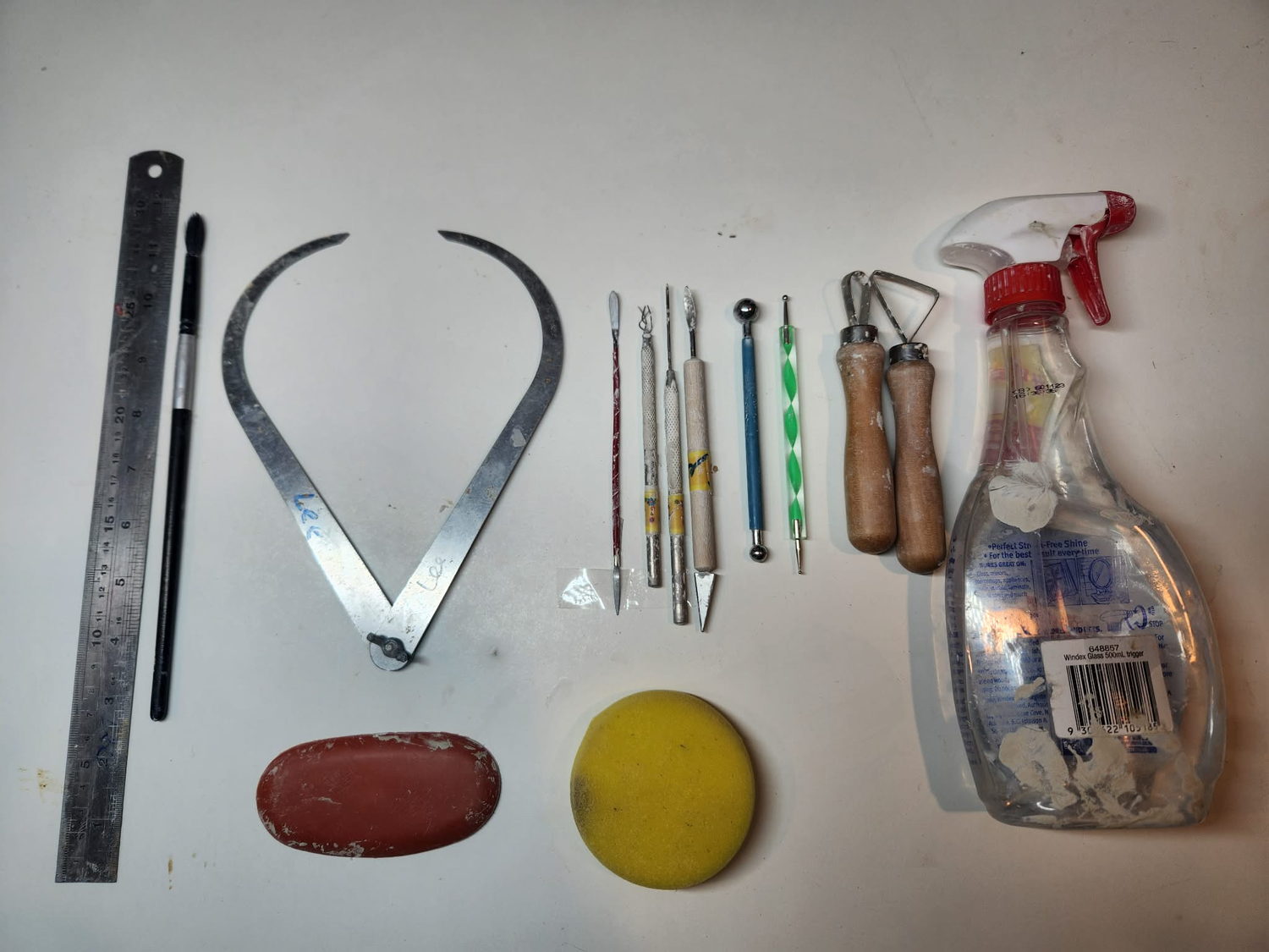 The minimum set of sculpting tools laid out on a white bench &mdash; a steel ruler, a soft brush, stainless steel calipers, fine drawing and eye tools, a ball tool, two wire loop tools, a red kidney rib, a round yellow sponge and a spray bottle